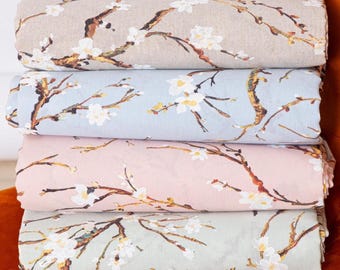 May include: A stack of four folded fabric pieces in various pastel colors: beige, light blue, pink, and mint green. Each piece features a repeating pattern of white blossoms and brown branches, creating a floral design. The fabrics are stacked on an orange surface.