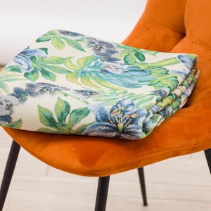 Può includere: Una coperta floreale piegata con sfondo crema, con disegni botanici blu e verdi. La coperta è drappeggiata su una sedia arancione con gambe nere, creando un contrasto vibrante.