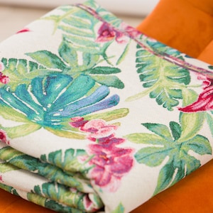 May include: Folded white blanket with a vibrant tropical floral pattern. The design features large green leaves, turquoise accents, and pink flowers. The blanket is folded, revealing the detailed print and texture of the fabric.