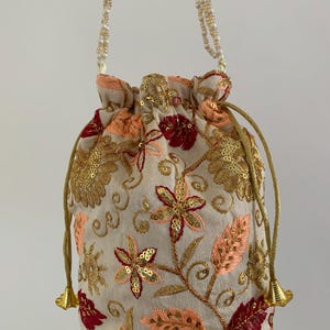 May include: A cream drawstring bag with detailed floral embroidery in gold, red, and peach. It has a beaded strap and gold drawstring cords. This ornate bag is a stylish accessory.