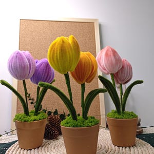 May include: Four potted faux tulip plants with green stems and fuzzy fabric flowers in shades of purple, yellow, orange, and pink. The pots are brown terracotta and are sitting on a woven straw mat.