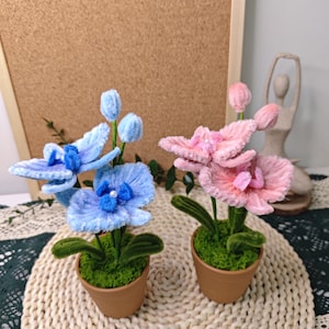 May include: Two potted faux orchid plants, one with blue flowers and one with pink flowers. The plants are in brown pots with green moss and are sitting on a woven straw mat.