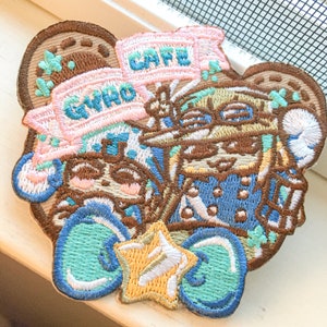 May include: Embroidered patch featuring a cartoon character with a cafe hat, glasses, and a blue and white patterned shirt. The patch also includes a star, hearts, and the text "GWHO CAFE".