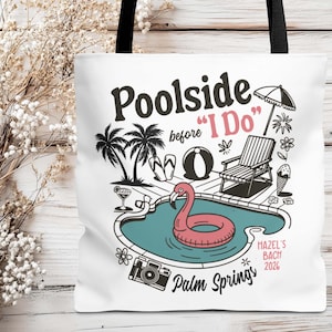 Könnte beinhalten: Weiße Tragetasche mit schwarzem Griff und Pool-Grafik. Das Design zeigt einen Pool mit Flamingo-Schwimmer, Palmen und den Texten "Poolside before I Do" und "Palm Springs 2026".