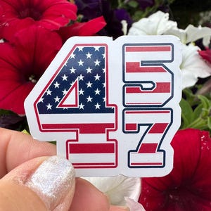 May include: A sticker featuring the numbers "47" in a patriotic design. The number "4" is filled with a blue background and white stars, while the "7" has red and white stripes. The sticker has a white border and is set against a floral background.