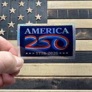 May include: A rectangular sticker with the word "AMERICA" in blue and red, with the number "250" in a swirling design. The sticker also includes the years "1776-2026" and is held in front of a wooden American flag.