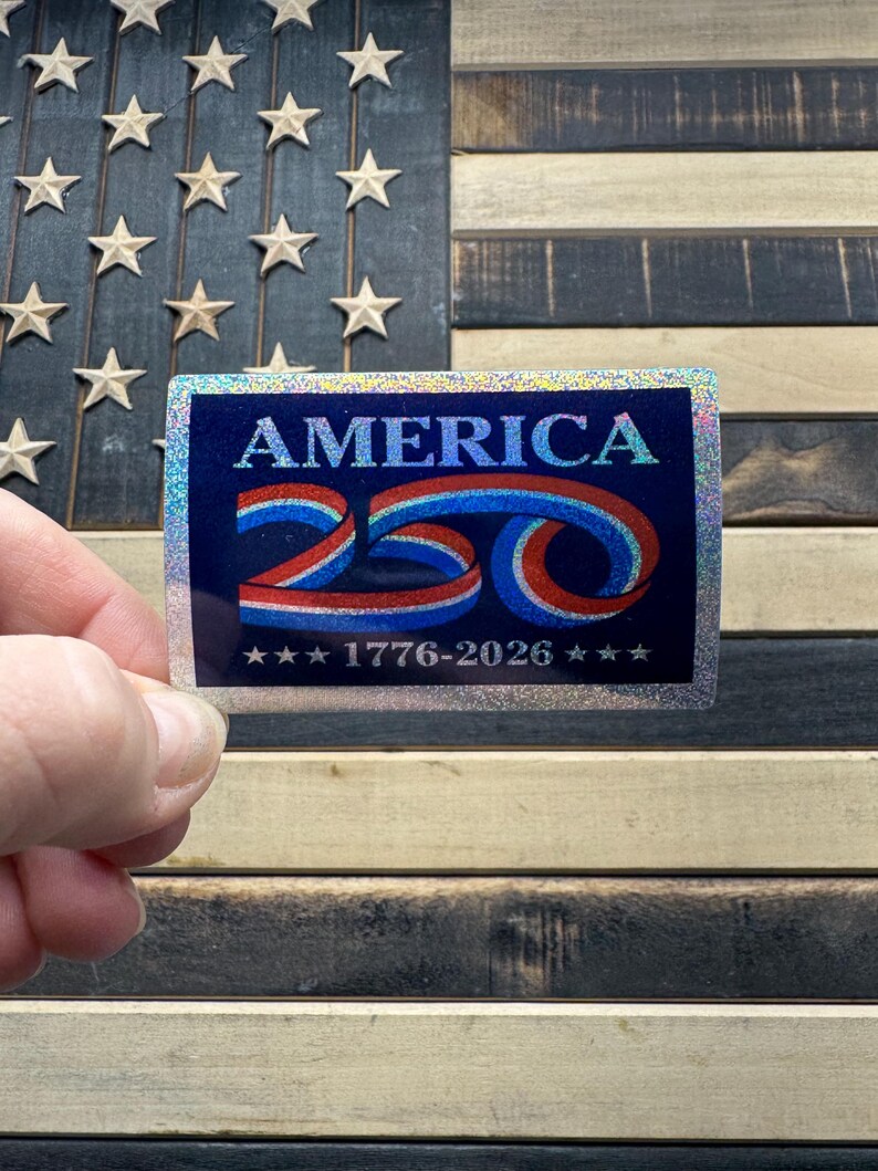 May include: A rectangular sticker with the word "AMERICA" in blue and red, with the number "250" in a swirling design. The sticker also includes the years "1776-2026" and is held in front of a wooden American flag.