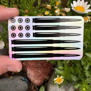 May include: A sticker with a black and white image of an American flag made of bullet casings. The sticker has a holographic iridescent background.