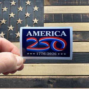 May include: A rectangular sticker with the word "AMERICA" in white capital letters over a red and blue ribbon design. Below the ribbon is the number "250" in red and blue, and the text "1776-2026" with stars. The sticker is held in front of a wooden American flag.