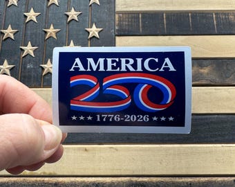 May include: A rectangular sticker with the word "AMERICA" in white capital letters over a red and blue ribbon design. Below the ribbon is the number "250" in red and blue, and the text "1776-2026" with stars. The sticker is held in front of a wooden American flag.