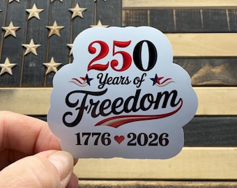 May include: A white sticker with the text "250 Years of Freedom" in red and black, with the years 1776 and 2026. The sticker is held in front of a wooden American flag.