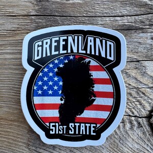 May include: A sticker with a white border and the word "GREENLAND" in bold, white letters on a black background. The design includes a silhouette of Greenland over an American flag and the text "51st STATE".
