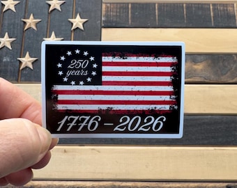 May include: A rectangular sticker featuring a stylized American flag design. The flag has red and white stripes with a black field of stars. The text "250 years" is above the flag, and "1776-2026" is below. The sticker is held by a hand.