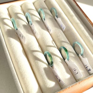 Könnte beinhalten: Eine Sammlung von Jade-Ringen in einer Vitrine. Die Ringe haben eine durchscheinende grüne Farbe und sind mit der Nummer "16.8" oder "16.9" auf kleinen weißen Etiketten versehen. Das Gehäuse hat einen hellfarbigen, gepolsterten Innenraum.