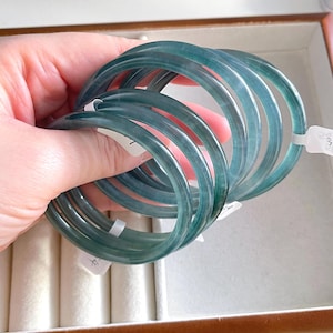May include: A collection of translucent, teal-colored jade bangles. The smooth, circular bracelets are held in a hand, showcasing their polished surface and varying shades of green and blue. The bangles are displayed in a jewelry box.