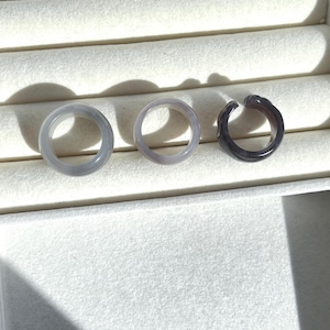 May include: Three rings in varying shades of gray, displayed on a white, textured surface. The rings are circular, with a smooth, polished finish. The first two rings are a light gray, while the third is a darker, charcoal gray.