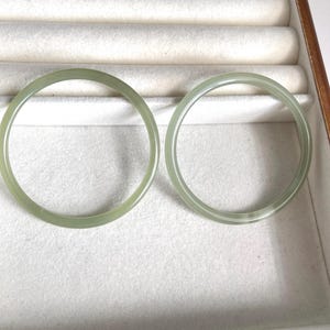 May include: Two pale green jade bangles are shown in a jewellery box. The bangles are circular, with a smooth, polished surface. The jewellery box has a light-coloured interior and a wooden frame.