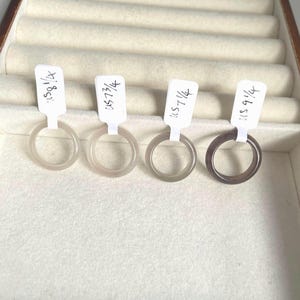 Könnte beinhalten: Vier durchscheinende Ringe in Grau- und Brauntönen werden in einer Schmuckschatulle präsentiert. Jeder Ring hat ein weißes Etikett mit handschriftlichem Text. Die Ringe variieren in der Farbe von hellgrau bis zu einem tiefen Braun.