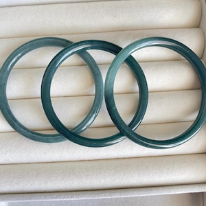May include: Three teal and green jade bangles are shown. The smooth, circular bracelets are arranged in a row, highlighting their polished surfaces. The bangles are displayed against a neutral, off-white background, creating a simple presentation.