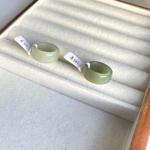 May include: Two pale green jade rings are presented in a jewellery box. Each ring has a smooth, polished surface and a rounded shape. The rings are labelled with small white tags. The jewellery box has a light-coloured, felt-lined interior.