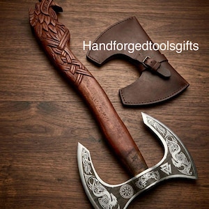 Puede incluir: Hacha artesanal con mango de madera y hoja de metal con intrincados diseños grabados. El mango está tallado con un detallado motivo de pájaro. Se incluye una funda de cuero marrón con hebilla. El texto "Handforgedtoolsgifts" es visible.