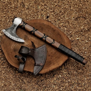 Puede incluir: Un hacha vikinga artesanal con un mango de madera envuelto en cuero negro. La cabeza del hacha es plateada con runas grabadas. Una funda de cuero protege la cabeza del hacha. El hacha descansa sobre una losa de madera.