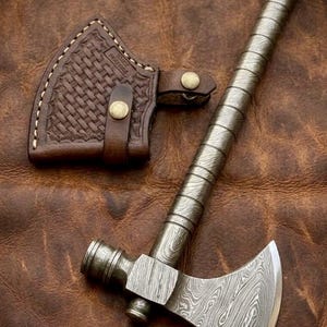 Könnte beinhalten: Ein Damaststahl-Beil mit einem langen, spiralförmigen Metallgriff und Hammerkopf, begleitet von einer braunen Lederscheide mit Korbgeflechtmuster, Messingdruckknöpfen und dem Text Handforgedtoolsgifts, der auf der Klinge sichtbar ist.