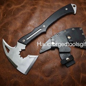 Puede incluir: Una hacha de metal plateado con mango negro y un diseño recortado cerca de la hoja, acompañada de una funda de cuero negro con broches plateados. El texto Handforgedtoolsgifts está impreso en blanco sobre la imagen.