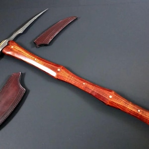 Könnte beinhalten: Eine handgefertigte Spitzhacke mit Holzgriff und Stahlklinge. Die Spitzhacke hat eine gebogene Klinge und ein spitzes Ende. Zwei Lederscheiden sind im Lieferumfang enthalten. Der Griff ist rotbraun mit kleinen Messingakzenten.