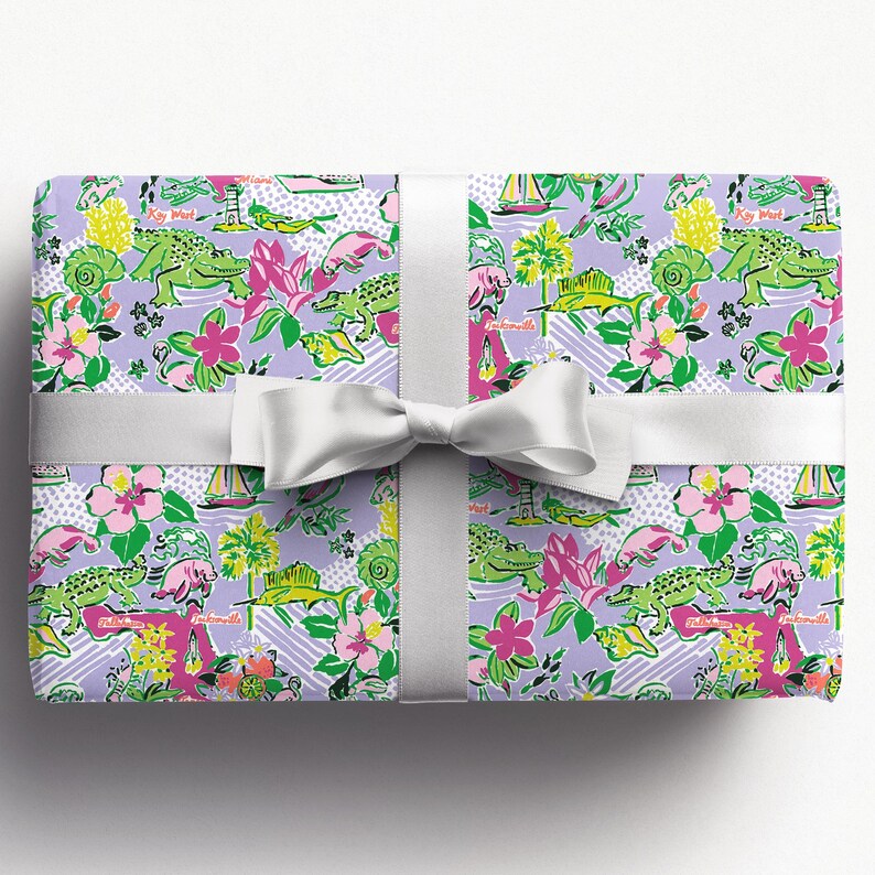 a gift wrapped in vibrant Florida-themed wrapping paper with playful illustrations of alligators, palm trees, and tropical flowers on a lavender background. A crisp white ribbon tied into a bow adds an elegant touch to the lively gift wrap.