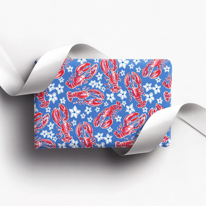 a gift wrapped in vibrant, preppy wrapping paper featuring bold red lobsters and white flowers on a blue background. The lively design is complemented by a crisp white ribbon, adding a touch of elegance to the playful gift wrap.