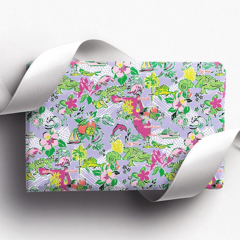 a gift wrapped in vibrant Florida-themed wrapping paper with playful illustrations of alligators, palm trees, and tropical flowers on a lavender background. A crisp white ribbon tied into a bow adds an elegant touch to the lively gift wrap.