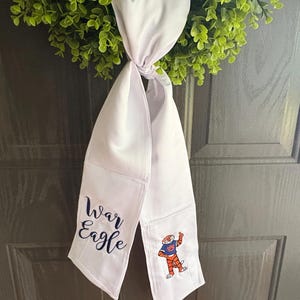 May include: White scarf with the words "War Eagle" in navy blue script and a cartoon tiger mascot. The scarf is hanging on a green wreath. The scarf is made of a soft, white fabric.