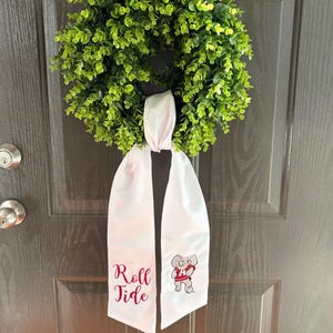 Könnte beinhalten: Ein grüner, blättriger Kranz hängt an einer dunkelbraunen Tür. Eine weiße Schärpe mit den Worten "Roll Tide" in burgunderfarbener Schrift und einer Cartoon-Elefanten-Grafik ist am Kranz befestigt. Die Tür hat einen silbernen Türknauf.