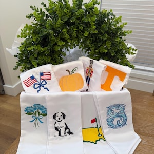 May include: A basket filled with white satin sashes, each embroidered with a unique design. Designs include an American flag, a pumpkin, a nutcracker, the letter "T", blue flowers, a dog, a golf course, and the letter "S". A green wreath sits behind the sashes.
