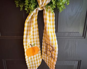 May include: A decorative wreath with a green leafy base and a yellow and white gingham ribbon. The ribbon features an embroidered orange pumpkin and the letter "R". The wreath is hanging on a dark grey door.