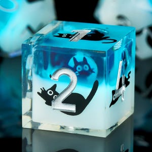 Könnte beinhalten: Ein blauer und weißer Würfel aus Harz mit einem schwarzen Katzenmotiv und der Zahl 2 auf einer Seite. Der Würfel ist mit einer klaren Flüssigkeit gefüllt und hat einen glitzernden Effekt.