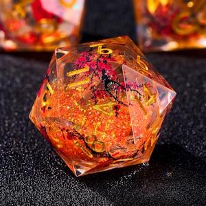 May include: A clear, multi-sided die with gold numbering and an interior design featuring a red and black floral pattern. The die is filled with orange glitter, creating a sparkling effect. The overall theme is fantasy gaming.