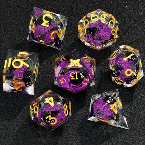 May include: Set of seven purple and gold dice with black cat and number designs. The dice are made of clear plastic with glitter embedded in the plastic.