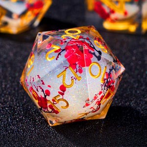 Op de afbeelding: Een veelzijdige, heldere harsdobbelsteen met gouden cijfers en rode bloeminsluitsels. De dobbelsteen heeft een glinsterend, wit interieur en is ontworpen voor tabletop games. De cijfers zijn goudkleurig.