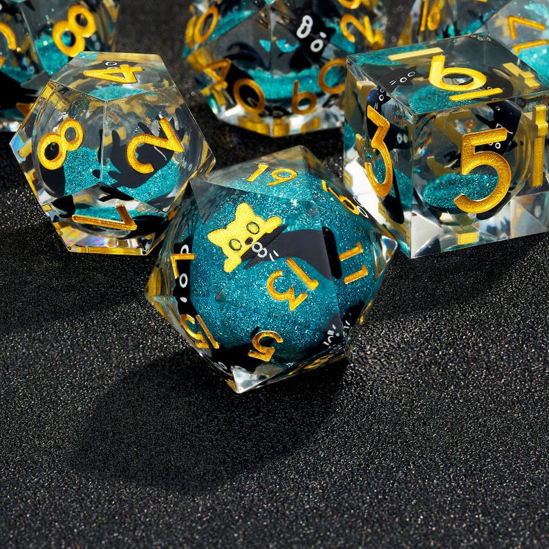 May include: A set of teal and gold dice with black numbers and a black cat graphic on one die. The dice are made of clear plastic with a glittery finish.