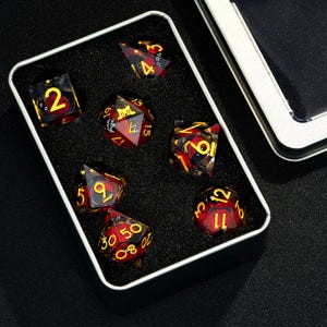 May include: A set of seven red and gold dice with black numbers in a black velvet lined metal box. The dice are translucent and have a swirl pattern.