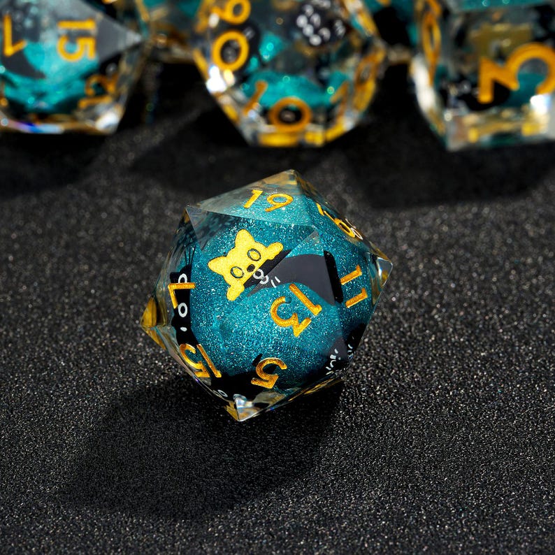 May include: A teal and gold d20 dice with a black and white illustration of a cat on one side. The dice has a glittery finish.