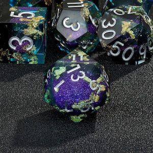 May include: A set of six iridescent dice with gold flecks. The dice are a deep purple and teal color with silver numbers. The dice are on a black surface.