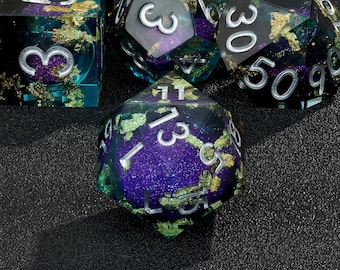 Peut inclure: Un ensemble de six dés irisés avec des paillettes dorées. Les dés sont d'une couleur violet foncé et turquoise avec des chiffres argentés. Les dés sont sur une surface noire.