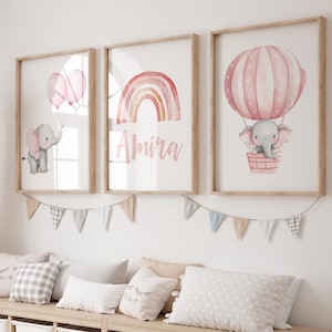 May include: Three framed watercolor prints with a nursery theme. One print features an elephant holding balloons, another a rainbow with the name "Amira", and the third an elephant in a hot air balloon. A wooden storage unit with white bins and pillows is below.