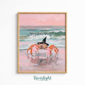 May include: A framed art print featuring an orange crab wearing a black pointed witch hat on a sandy beach. The background shows a soft pink sky and teal ocean waves crashing on the shore. The text Riverlight PRINTS is visible at the bottom.