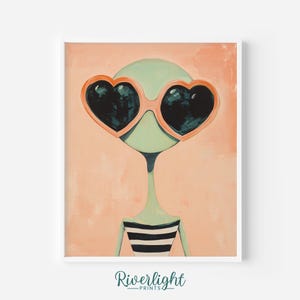 May include: A framed art print featuring a whimsical alien figure with a light green head and body, wearing heart-shaped sunglasses with black lenses. The alien is dressed in a black and white striped top, set against a peach-colored background.