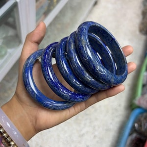 以下が含まれることがあります： 手には、磨かれた濃い青色の宝石バングルが5つ重ねられています。 バングルは滑らかで光沢のある仕上がりで、色と質感に微妙な変化があります。 ブレスレットは豊かで鮮やかな青色で、明るい色合いが少しあります。
