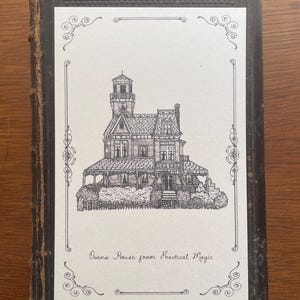 Può includere: Un disegno a penna in bianco e nero di una casa vittoriana, intitolato "Owens House from Practical Magic", è incorniciato da un bordo decorativo. L'opera d'arte è presentata su una copertina di libro vintage con una finitura in pelle marrone scuro e usurata.